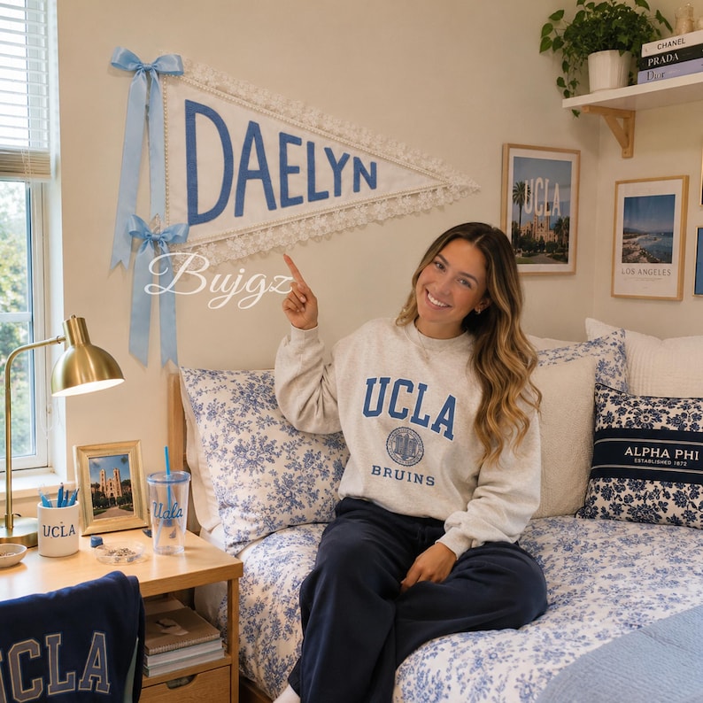 May include: A triangular white felt pennant featuring the name DAELYN in blue block letters, trimmed with white lace and adorned with two light blue satin ribbons. The pennant is displayed on a wall above a bed with blue and white floral bedding.