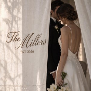 May include: A white linen curtain featuring the brown printed text The Millers EST 2026. Behind the curtain, a person in a white backless wedding dress with thin straps and a row of buttons stands next to a person in a black tuxedo with a boutonniere.