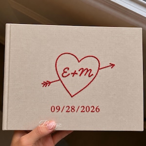 Puede incluir: Un álbum de fotos rectangular cubierto de tela beige con bordados en rojo. El diseño incluye un corazón atravesado por una flecha con las letras E+M en su interior, y la fecha 09/28/2026 bordada en hilo rojo debajo del corazón.