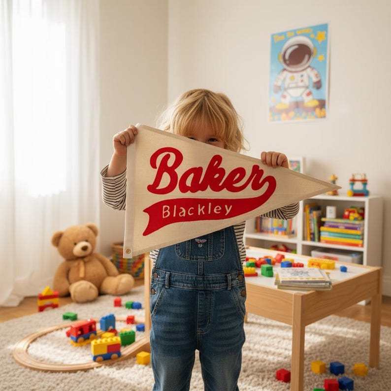 K&ouml;nnte beinhalten: Ein cremefarbenes Wimpel mit dem Namen "Baker" in roter Schrift und "Blackley" in kleinerer Schrift. Der Wimpel wird von einem Kind in einem Raum mit Spielzeug und einem Teddyb&auml;r gehalten.