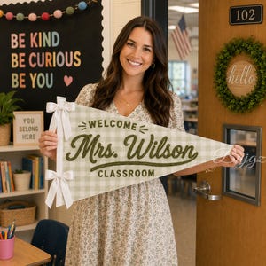 Peut inclure: Un fanion triangulaire en tissu avec un motif vichy vert clair et blanc. Le texte WELCOME est brodé en vert foncé, suivi du nom Mrs. Wilson en écriture cursive, et du mot CLASSROOM. Deux rubans blancs sont attachés sur le côté du fanion.