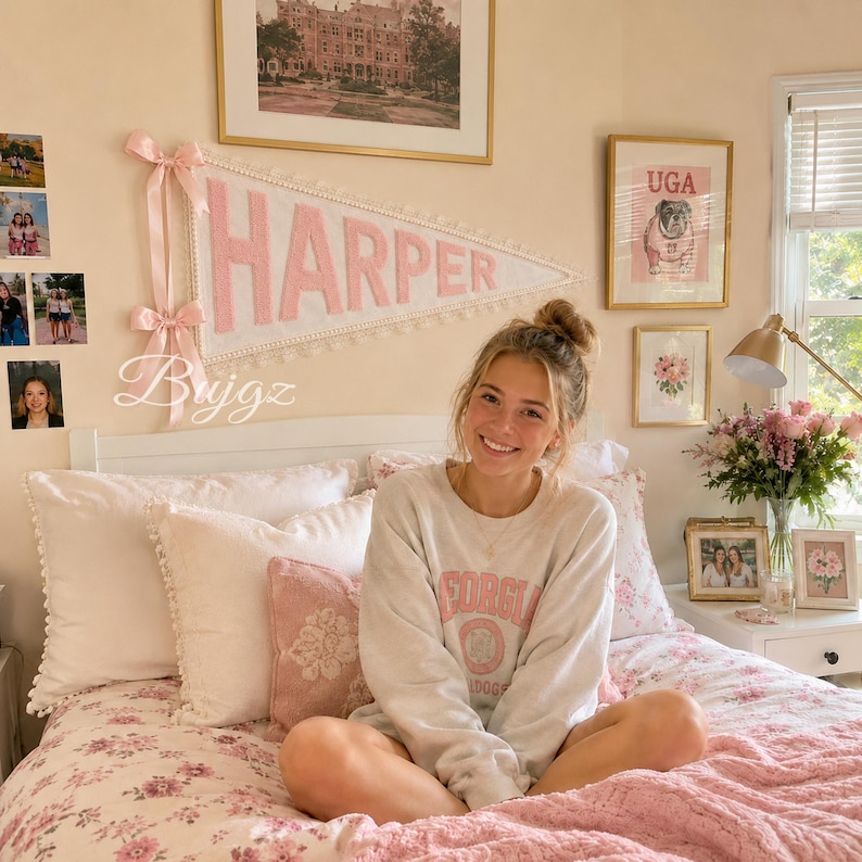 May include: A white felt pennant banner with the name HARPER in large pink letters, featuring a decorative lace trim edge and two pink ribbon bows on the left side. The banner is mounted on a wall above a bed with pink and white bedding.