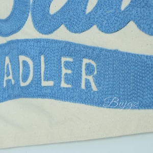 K&ouml;nnte beinhalten: Nahaufnahme einer Canvas-Tasche mit dem Namen "ADLER" in Wei&szlig; auf einem hellblauen Banner gestickt. Die Oberseite der Tasche zeigt ein blaues Wellenmuster. Der Hintergrund ist cremewei&szlig;.