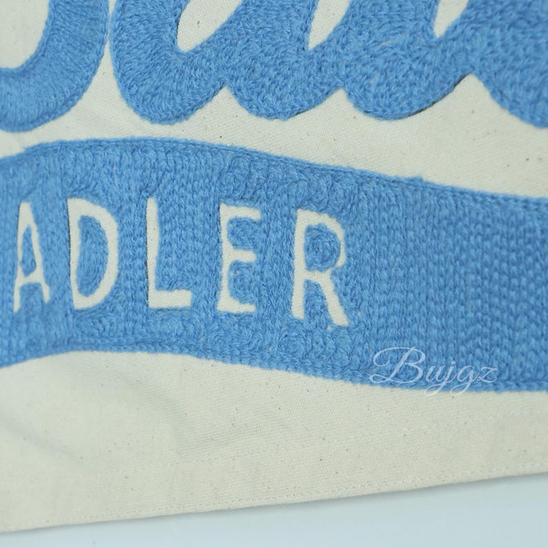 K&ouml;nnte beinhalten: Nahaufnahme einer Canvas-Tasche mit dem Namen "ADLER" in Wei&szlig; auf einem hellblauen Banner gestickt. Die Oberseite der Tasche zeigt ein blaues Wellenmuster. Der Hintergrund ist cremewei&szlig;.