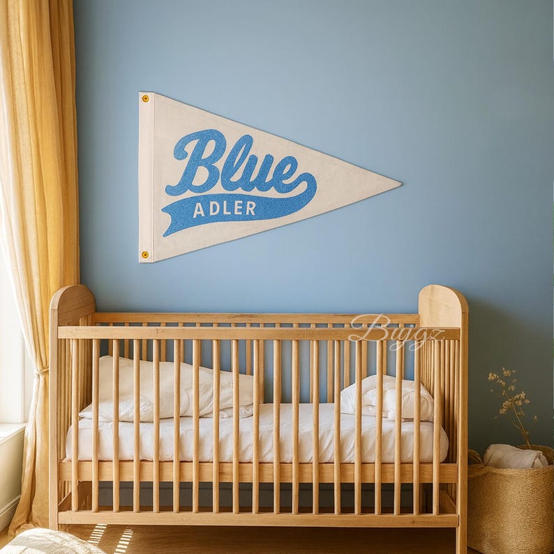 K&ouml;nnte beinhalten: Ein cremefarbener Wimpel mit dem Wort "Blue" in blauer Schrift und "ADLER" in blauer serifenloser Schrift. Der Wimpel h&auml;ngt an einer hellblauen Wand &uuml;ber einem Holzbettchen.
