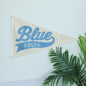 K&ouml;nnte beinhalten: Cremefarbener Wimpel mit dem Wort "Blue" in hellblauer Schrift und "ADLER" darunter. Die dreieckige Flagge hat Metall&ouml;sen zum Aufh&auml;ngen. Gr&uuml;ne Pflanzenbl&auml;tter sind im Hintergrund zu sehen.