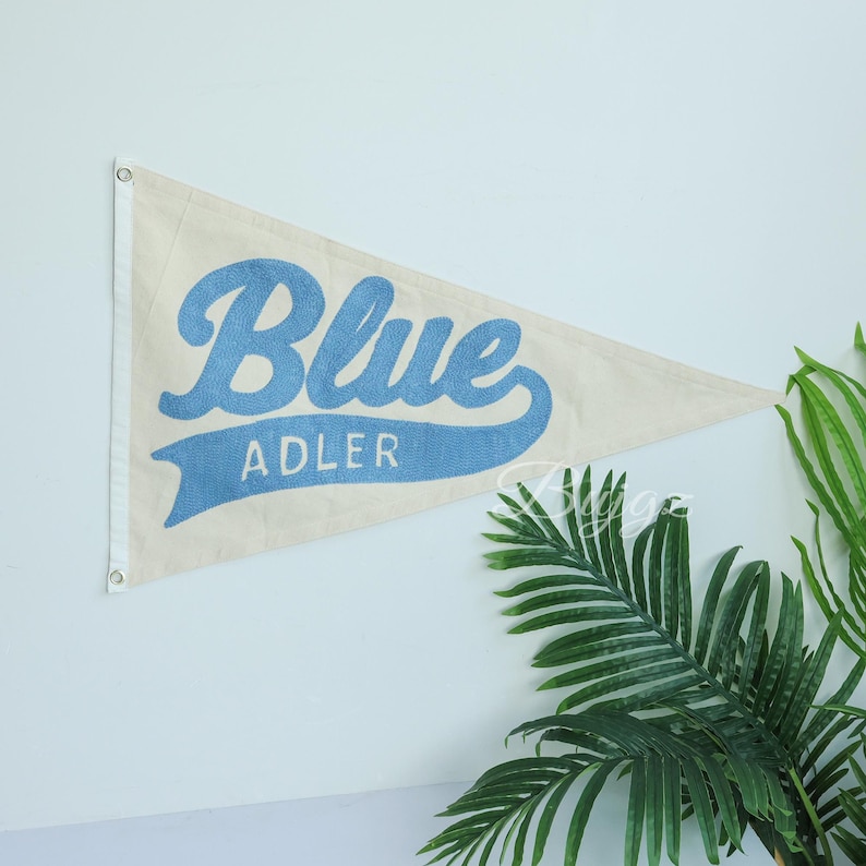 K&ouml;nnte beinhalten: Cremefarbener Wimpel mit dem Wort "Blue" in hellblauer Schrift und "ADLER" darunter. Die dreieckige Flagge hat Metall&ouml;sen zum Aufh&auml;ngen. Gr&uuml;ne Pflanzenbl&auml;tter sind im Hintergrund zu sehen.