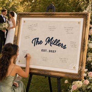 May include: A large rectangular guest book sign in an ornate gold frame featuring a cream-colored fabric center with metal grommets. The text reads The Millers EST. 2027 in dark blue script and block letters, surrounded by handwritten guest messages.