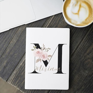 Könnte beinhalten: Weißes Notizbuch mit einem großen schwarzen Buchstaben "M", verziert mit rosa Rosen und Grün. Der Name "Miriam" ist in einer Schreibschrift unter dem Buchstaben geschrieben. Ein Laptop und eine Kaffeetasse sind im Hintergrund.