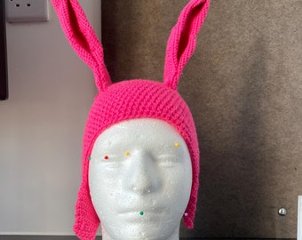 May include: A bright pink knitted hat featuring two long, upright rabbit ears. The hat is constructed with a textured stitch pattern and includes ear flaps that extend down the sides. It is displayed on a white foam mannequin head.
