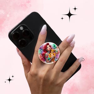 May include: A black smartphone with a round pop socket featuring a colorful illustration of a woman with red hair and tattoos holding a lion cub. The background is a watercolor design with pink, green, and purple hues.