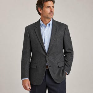 May include: A dark gray blazer with a subtle micro-check pattern, featuring notched lapels, two front buttons, and flap pockets. The blazer is worn over a light blue button-down shirt, paired with dark navy trousers, creating a structured look.
