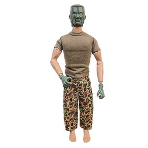 May include: A vintage action figure of a soldier, featuring a green camouflage painted face and olive drab t-shirt. The figure wears camouflage pants with a brown, green, and tan pattern. The figure has articulated limbs and green gloves.