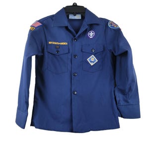 May include: A navy blue Boy Scouts of America uniform shirt with long sleeves. The shirt has two chest pockets and several patches, including an American flag and a Boy Scout emblem. The text "BOY SCOUTS OF AMERICA" is embroidered above the pockets.