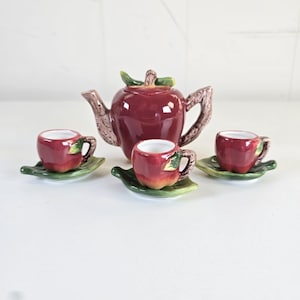 May include: A ceramic tea set featuring a teapot and three cups with matching saucers, all shaped like red apples. The teapot has a brown branch-like handle and spout, while each cup sits on a green leaf-shaped saucer with a small green leaf accent.