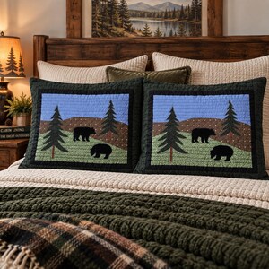 May include: Two rectangular quilted pillow shams featuring a central graphic of two black bears in a forest landscape with green hills, dark green pine trees, and a light blue sky, all framed by a thick dark green border with a stitched texture.