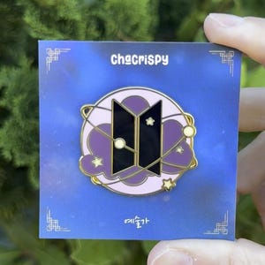 Op de afbeelding: Vierkante pin met een paars en goudkleurig ontwerp. De pin heeft de tekst "ChaCrispy" en Koreaanse tekst. De pin heeft een zwart en goudkleurig logo met een ster- en planeetontwerp.