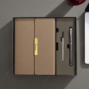 May include: A brown leather-bound journal with a gold nameplate that reads "Susan" is in a black gift box with a silver pen and a black pen.