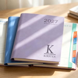 May include: A lavender 2027 schedule notebook with a large letter K and the name KIRSTEN printed on the front cover. The notebook is layered over a blue notebook and an open planner with colourful tabbed dividers on the right side of the page.