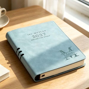 Könnte beinhalten: Ein hellblauer Planer mit matter Oberfläche, der den Text 365 MY PLAN 2027 WRITING FOR MYSELF sowie ein Monogramm mit dem Namen Meiti trägt. Das rechteckige Buch hat genähte Kanten und einen sichtbaren Buchrücken an der linken Seite.