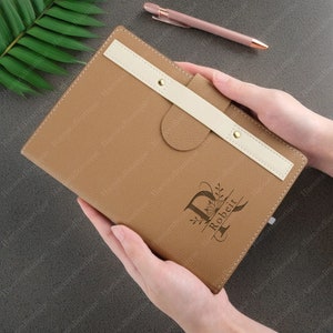 May include: A brown leather journal with a white leather strap and a gold metal closure. The journal is personalised with the letter "R" and the name "Robert" in a decorative font.
