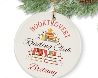 May include: A round, white ceramic ornament with the text "BOOKTROVERT Reading Club Britany" in burgundy and black script. The design includes illustrations of books and a star, with festive string lights. A gold string is attached for hanging.