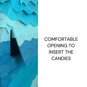 Puede incluir: Primer plano de una manualidad de papel azul y turquesa con el texto "COMFORTABLE OPENING TO INSERT THE CANDIES". La manualidad tiene una apariencia en capas y texturizada, lo que sugiere una pi&ntilde;ata o un art&iacute;culo similar.