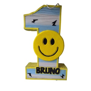 May include: A number one birthday decoration with a yellow smiley face and the name "BRUNO" in black block letters. The number is blue and white with yellow trim and lightning bolt accents. A white string is attached for hanging.