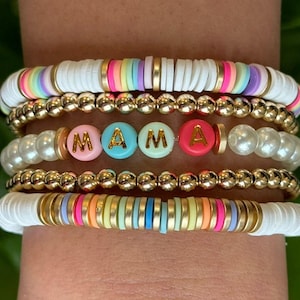 May include: A stack of five beaded bracelets featuring gold-tone metallic spheres, white faux pearls, and flat polymer clay discs in rainbow colours. The centre bracelet includes four round beads with the gold-coloured letters M, A, M, and A.