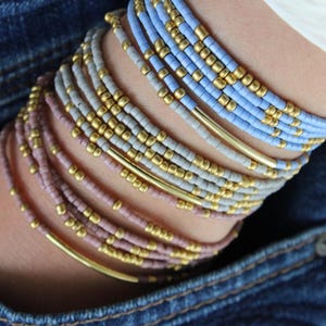 May include: A collection of beaded bracelets in shades of pink, gray, and blue, accented with gold-colored metal tubes and beads. The bracelets are stacked on a wrist, set against a denim background.