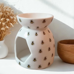 Peut inclure: Un brûleur d'huile en céramique blanche avec un motif d'abeilles. Le brûleur a un dessus rond pour l'huile et une grande ouverture à l'avant pour une bougie. Le motif d'abeilles présente des abeilles noires et jaunes. Un objet décoratif pour parfumer la maison.
