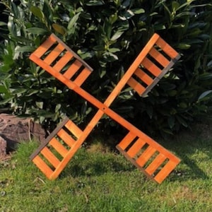 Könnte beinhalten: Eine Windmühle aus Holz mit vier orangefarbenen Flügeln. Die Flügel sind kreuzförmig angeordnet und haben einen dunkelbraunen Rand. Die Windmühle steht auf einem grünen Rasen.