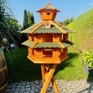 Könnte beinhalten: Ein mehrstöckiges Vogelhaus aus Holz mit einem grünen Schindeldach und einer natürlichen Holzoberfläche. Das Vogelhaus ist auf einem Holzständer montiert und von grünem Gras und Laub umgeben. Das Vogelhaus hat mehrere Öffnungen.