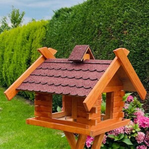 Könnte beinhalten: Ein Vogelhaus aus Holz mit einem roten Schindeldach. Der Futterspender hat zwei Fütterungsplätze und eine Plattform darunter. Der Futterspender steht auf einem hohen Holzständer.
