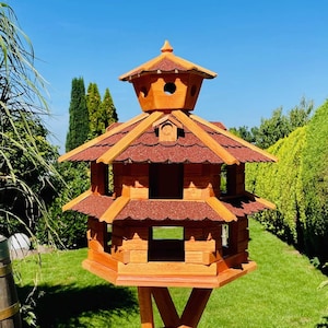 Könnte beinhalten: Ein Vogelhaus aus Holz mit mehrstöckigem Design und einem roten Schindeldach. Das Haus ist auf einem hohen Holzpfahl montiert.