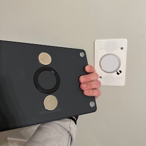 Puede incluir: Un dispositivo rectangular negro con tres almohadillas adhesivas redondas plateadas y un anillo negro en el centro. El dispositivo está siendo sostenido por una mano y se está colocando en una pared blanca.