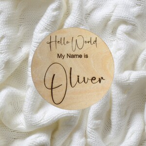 May include: A round wooden sign with the text "Hello World My Name is Oliver" engraved on it. The sign is on a white knitted blanket.