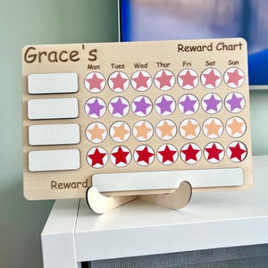 May include: Wooden reward chart with the name "Grace's" at the top. The chart features rows of star-shaped spaces in shades of pink, purple, orange, and red for tracking progress. Includes blank spaces for rewards and a wooden stand.