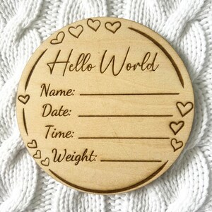 May include: A round wooden plaque with the text "Hello World" and spaces to fill in the baby's name, date, time, and weight. The plaque is decorated with small hearts around the edge.