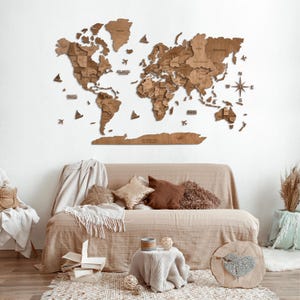 K&ouml;nnte beinhalten: Eine dekorative Weltkarte aus Holz an der Wand mit einem Kompass und den Namen der Ozeane. Die Karte ist an einer wei&szlig;en Wand &uuml;ber einem beigen Sofa mit Kissen und einer Decke angebracht.