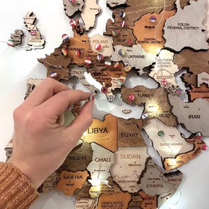 Può includere: Una mappa del mondo in legno con contorni di paesi e spilli con bandiera. La mappa è realizzata in varie tonalità di legno e include luci a stringa a LED. Una mano sta posizionando uno spillo con bandiera sulla mappa.