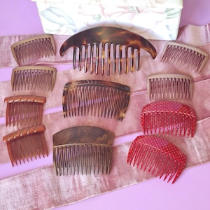 May include: A collection of vintage hair combs in various styles and colors, including tortoiseshell, brown, and red with white polka dots. The combs are arranged on a pink ribbon against a light purple background, suggesting a vintage or antique theme.