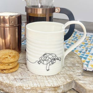 May include: White ceramic mug with a black line drawing of a turtle on the side. The mug has a ribbed design and a rounded handle.