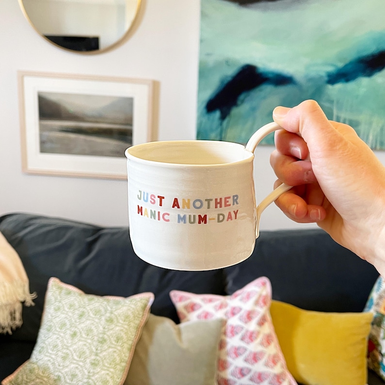 May include: White ceramic mug with the text "JUST ANOTHER MANIC MUM-DAY" in colorful letters. The mug is being held by a hand in front of a couch with pillows.