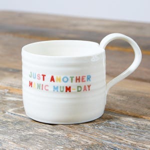 May include: White ceramic mug with a colorful inscription that reads "Just Another Manic Mum-Day".
