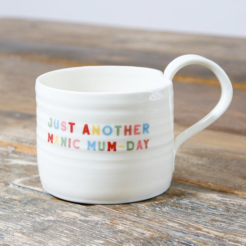 May include: White ceramic mug with a colorful inscription that reads "Just Another Manic Mum-Day".