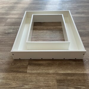 May include: A white rectangular frame with a smaller white rectangular frame inside. The frame is made of a smooth, white material and has a slightly raised edge. The frame is sitting on a wooden surface.