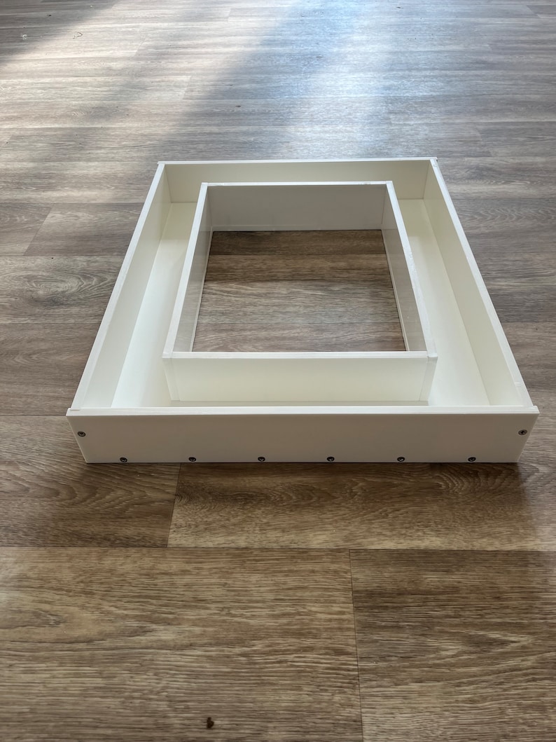 May include: A white rectangular frame with a smaller white rectangular frame inside. The frame is made of a smooth, white material and has a slightly raised edge. The frame is sitting on a wooden surface.