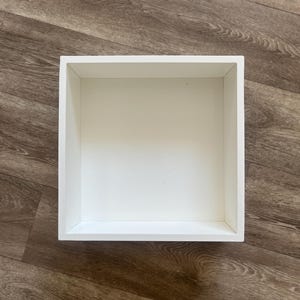 Peut inclure: Une étagère carrée en bois blanc. La boîte est vide et présente un design épuré et minimaliste. L'étagère est posée sur un sol en bois avec un motif de grain de bois brun.
