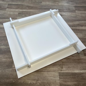 Op de afbeelding: Witte kunststoffraam voor een foto of kunstwerk. Het frame is gemaakt van vier stukken die in elkaar passen om een vierkante vorm te creëren. Het frame ligt op een wit oppervlak.